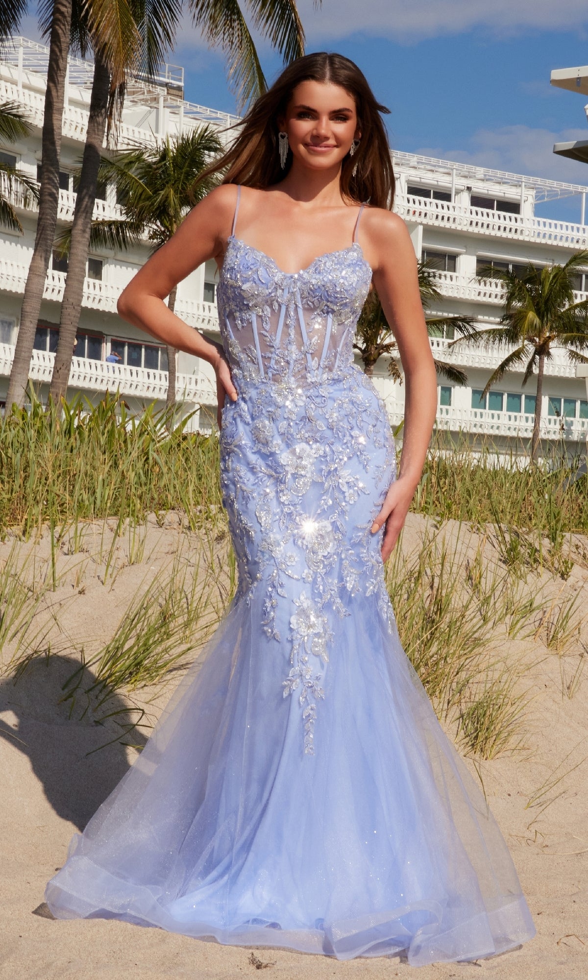 A woman in the Long Embroidered Prom Dress: Nox Anabel Q1541 stands on a sandy beach with grass, palm trees, and a white building in the background.